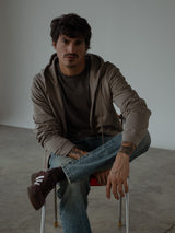 Modelo sentado en una silla, usando buzo unisex color mocca con chompa y cremallera, camiseta color café y jean azul clásico.
