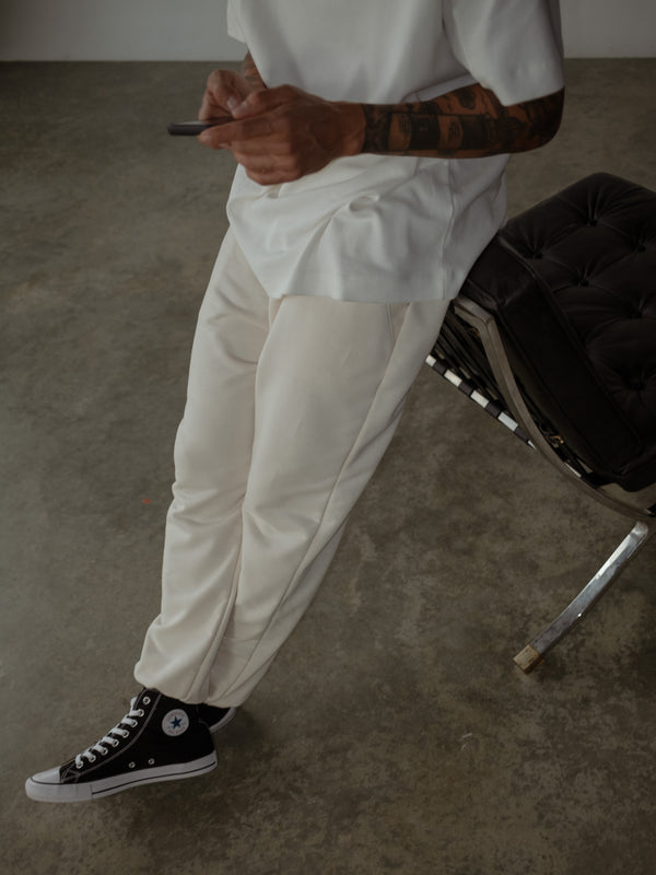 Primer plano de modelo sentado en una silla, usando jogger color crema con bota elástica y camiseta oversized color crudo, mientras usa un teléfono.