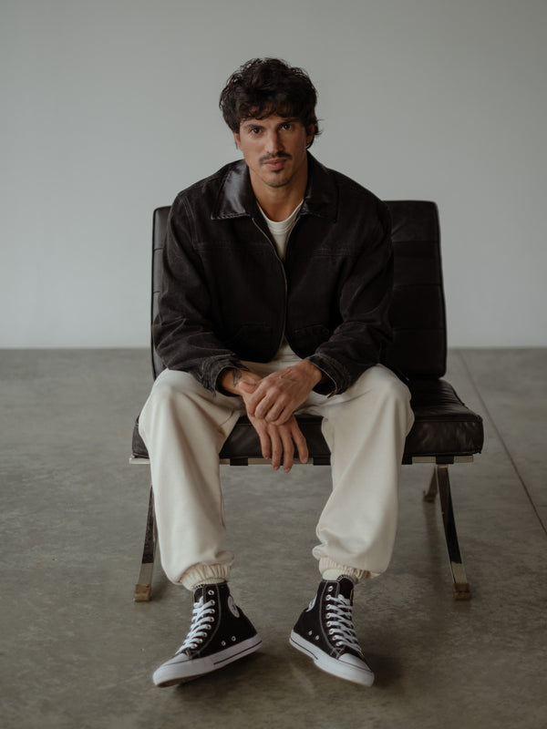 Modelo sentado en un sillón, usando jogger color crema con bota elástica, camiseta oversized color crudo y chaqueta de jean negra.