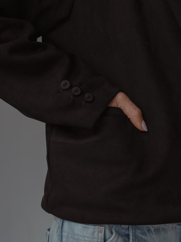 Primer plano de una mano en el bolsillo lateral de un blazer color negro, mostrando los botones del puño y un jean azul claro.