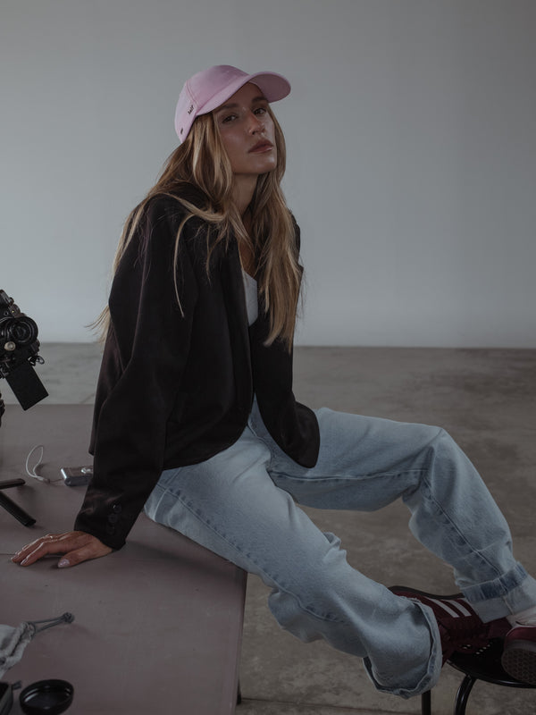 Modelo sentada en una superficie, usando un top asimétrico color crema, un jean azul claro, una gorra rosada y un blazer color negro abierto.