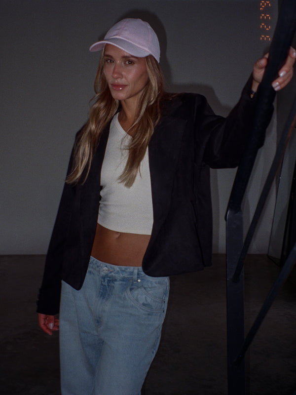 Modelo con gorra rosada, recostada en una baranda, usando un blazer color negro, un top de rib color crema y un jean azul claro.