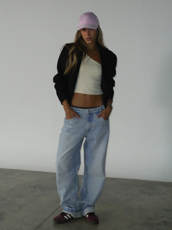 Modelo con gorra rosada y las manos en los bolsillos, usando un blazer color negro abierto, un top en rib color crema y un jean azul claro.