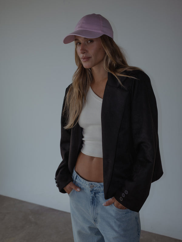 Modelo con las manos en los bolsillos, usando un blazer color negro, un top asimétrico en rib color crema, una gorra rosada y un jean azul claro.