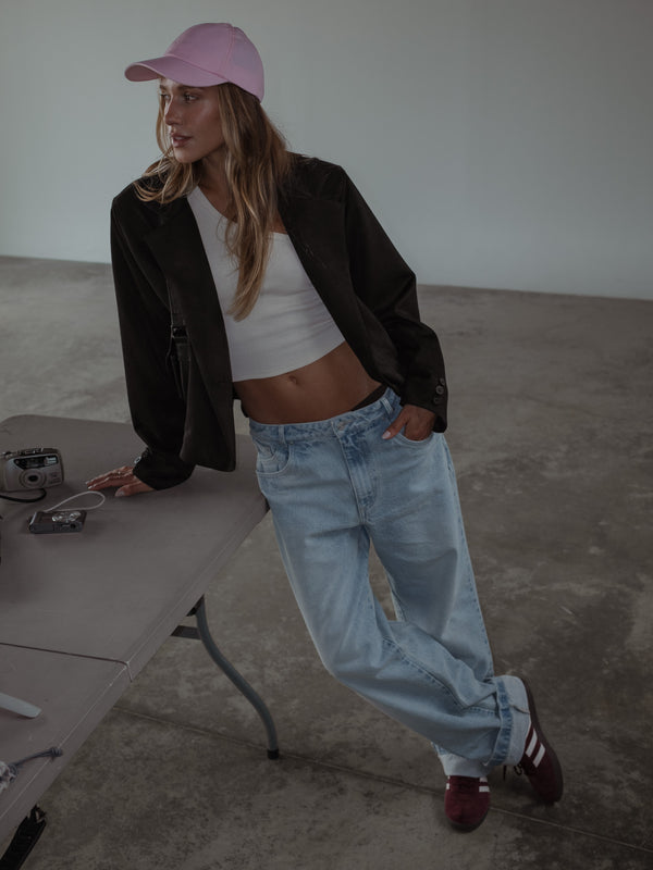 Modelo apoyada en una mesa, usando un jean azul claro, un top asimétrico en rib color crema, una gorra rosada y un blazer color negro.