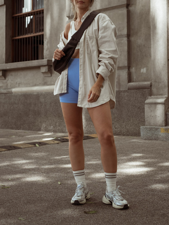Modelo usando un short azul, crop top blanco, camisa manga larga y bolso café