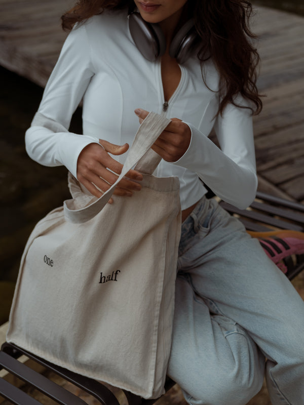Mujer sentada sosteniendo un bolso color crema con texto estampado color negro "one half"