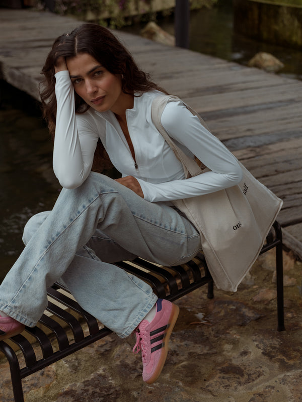 Mujer sentada de perfil con un bolso color crema con texto negro "one half" al hombro