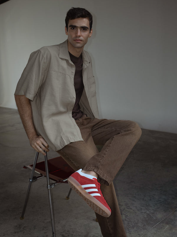 Modelo sentado en una silla metálica, usando una camisa manga corta color arena sobre una camiseta color café y jeans color café.