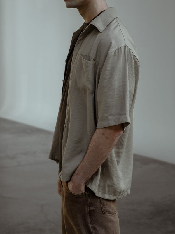 Modelo de perfil con una mano en el bolsillo de sus jeans color café, usando una camiseta color café bajo una camisa manga corta color arena.