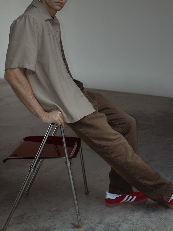 Modelo sentado de lado en una silla, usando jeans color café y una camisa manga corta color arena.