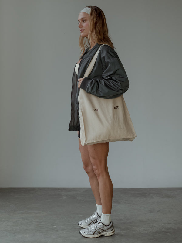 Modelo de perfil, sosteniendo tote bag color crema con el texto "one half" estampado, usando chaqueta tipo bomber color gris, top color crema, shorts negros y headband color crema.