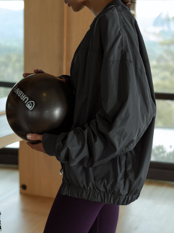 Modelo de perfil, sosteniendo balón, usando chaqueta tipo bomber color gris y leggings deportivos color morado.