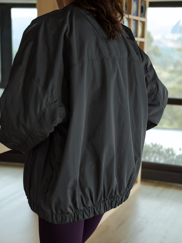 Modelo de espaldas, usando una chaqueta tipo bomber color gris y leggings deportivos color morado