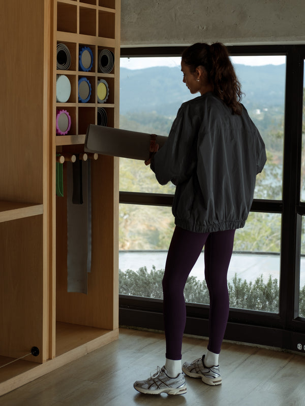 Modelo de perfil, sosteniendo una esterilla de yoga, usando una chaqueta tipo bomber color gris y leggings deportivos color morado.