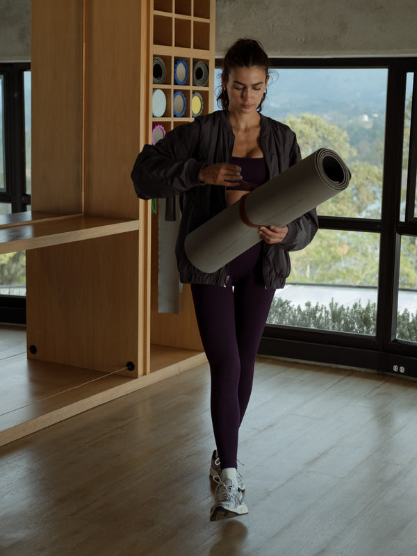 Modelo caminando, sosteniendo una esterilla de yoga, usando una chaqueta tipo bomber color gris, un top y leggings deportivos color morado.