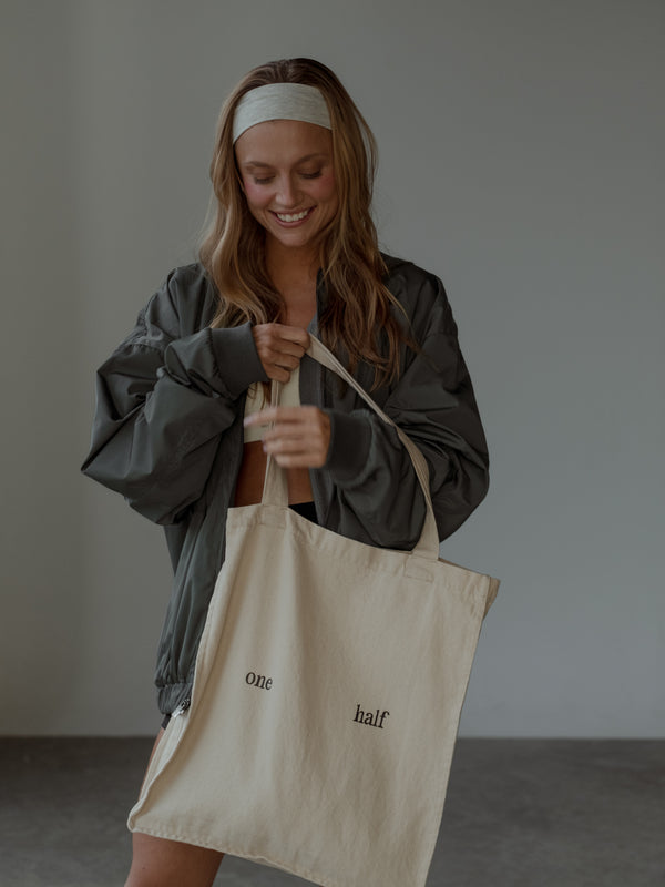 Modelo sonriendo, sosteniendo tote bag crema con el texto "one half" estampado, usando chaqueta tipo bomber color gris, top color crema, shorts negros y headband color crema.