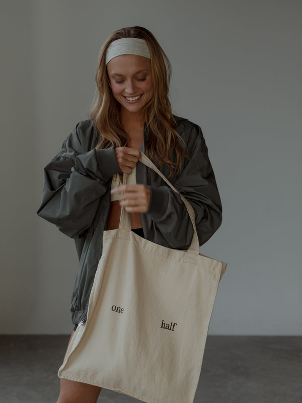 Modelo sonriendo, sosteniendo tote bag crema con el texto "one half" estampado, usando chaqueta tipo bomber color gris, top color crema, shorts negros y headband color crema.