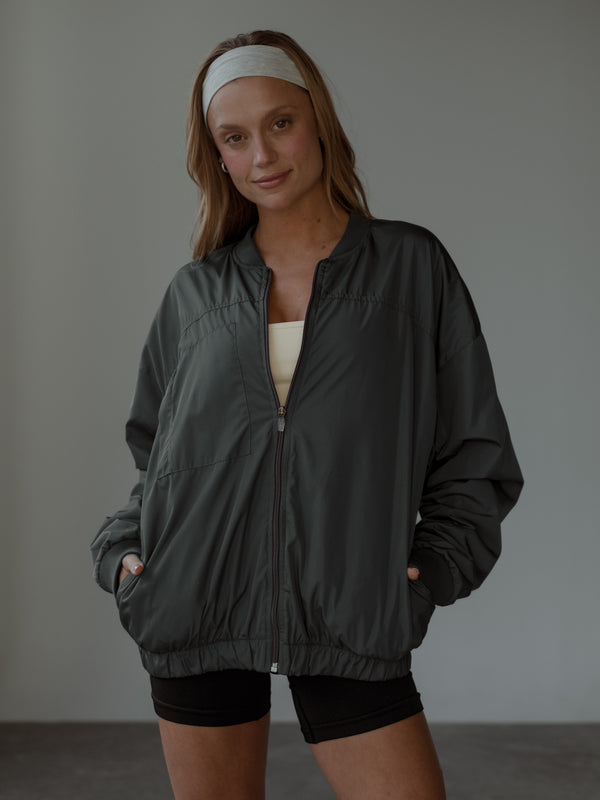 Modelo de frente, sonriendo, con las manos en los bolsillos, usando una chaqueta tipo bomber color gris, top color crema, shorts negros y headband color crema.