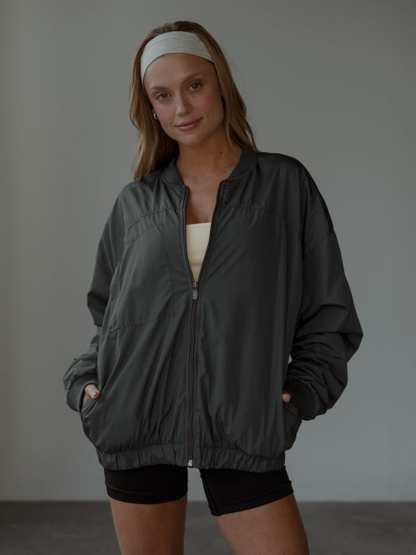 Modelo de frente, sonriendo, con las manos en los bolsillos, usando una chaqueta tipo bomber color gris, top color crema, shorts negros y headband color crema.