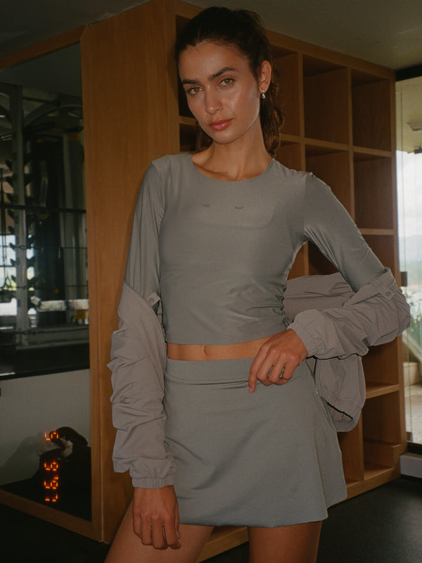 Modelo de pie con chaqueta gris sobre un hombro usando falda deportiva gris y top manga larga gris