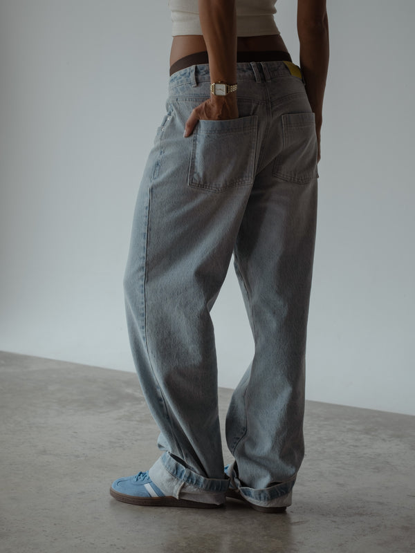 Plano medio de modelo de espalda con una mano en el bolsillo trasero, luciendo un jean bota recta color azul de tiro medio, un top corto blanco y ropa interior color café.