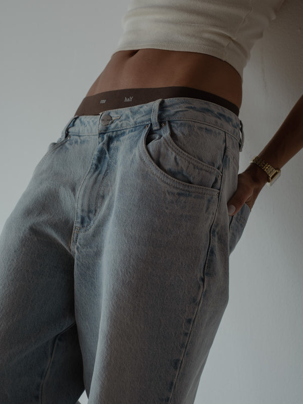 Primer plano de modelo con una mano en el bolsillo trasero de un jean color azul de tiro medio y doble bolsillo frontal, combinado con un top corto blanco y ropa interior negro.