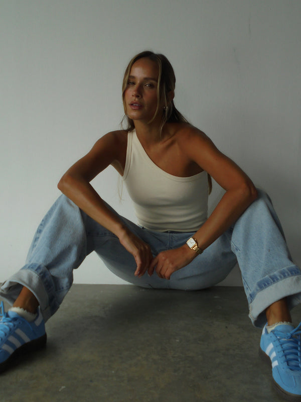 Modelo sentada en el suelo con las piernas extendidas, usando un top asimétrico color crema y un jean bota recta color azul de tiro medio y doble bolsillo frontal.