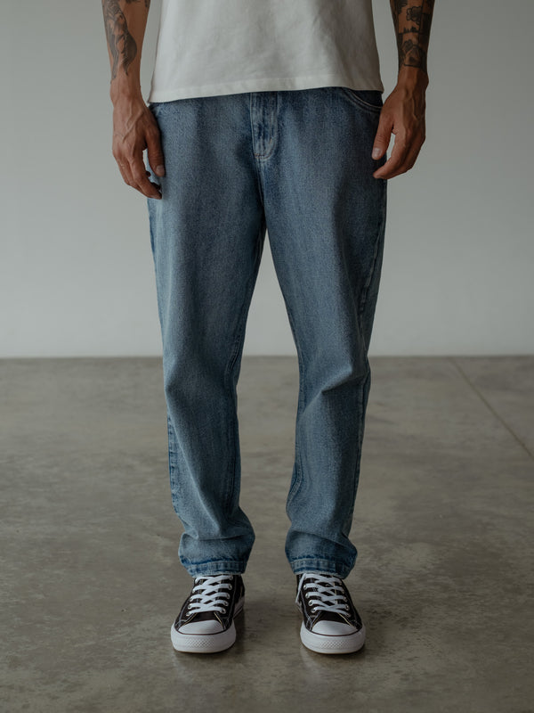 Primer plano de la parte inferior de modelo de pie, usando jean azul de bota recta con bolsillos frontales y camiseta oversized color crudo.