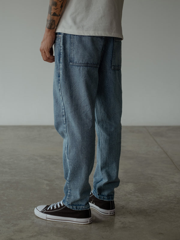 Primer plano posterior de modelo de pie, usando jean azul de silueta holgada con bolsillos y camiseta oversized color crudo.