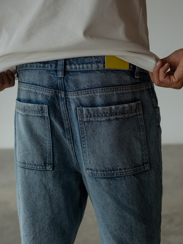 Primer plano posterior de modelo ajustando su camiseta color crudo, dejando ver la parte posterior del jean azul con bolsillos posteriores.