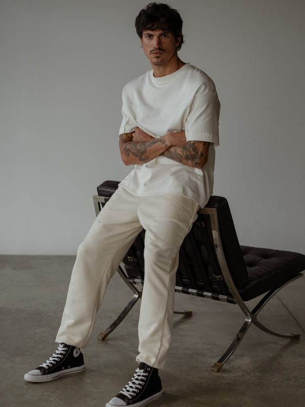 Modelo sentado en un sillón con los brazos cruzados, usando jogger color crema con bota elástica y camiseta oversized color crudo.
