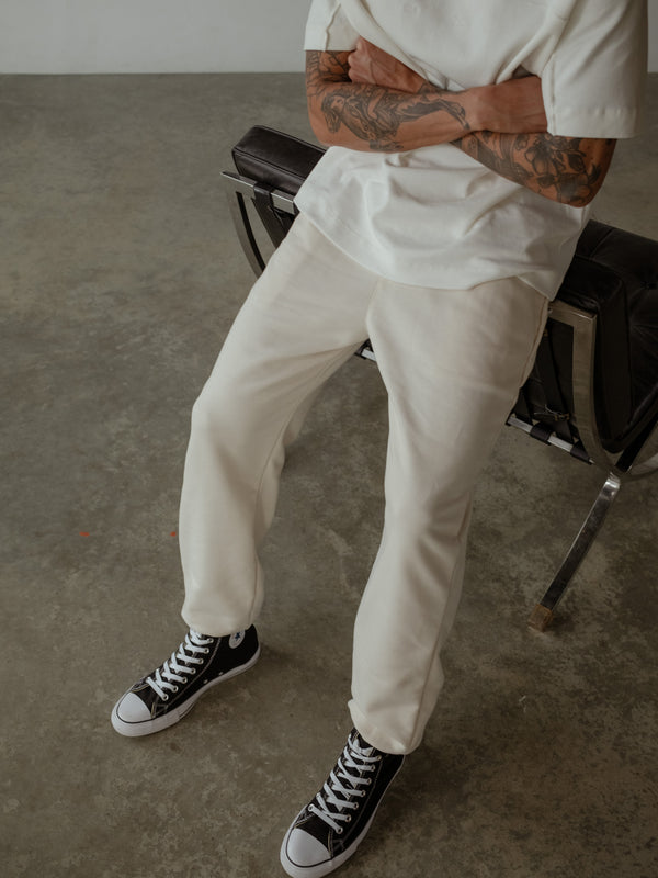 Primer plano de modelo sentado con los brazos cruzados, usando jogger color crema con bota elástica y camiseta oversized color crudo.