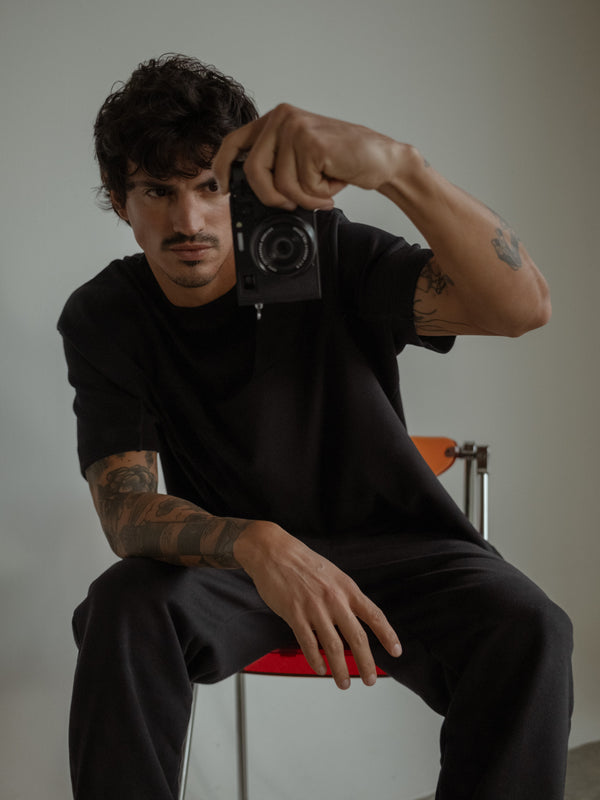 Modelo sentado en una silla, usando jogger color negro con bota elástica y camiseta oversized negra, mientras sostiene una cámara.