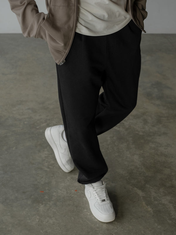 Primer plano inferior de modelo de pie, usando jogger color negro con bota elástica y bolsillos laterales, camiseta oversized blanca y buzo con cremallera café.