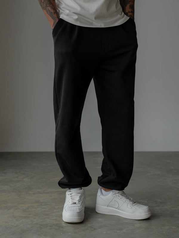 Primer plano inferior de modelo de pie con manos en los bolsillos de un jogger color negro con bota elástica y bolsillos laterales, y camiseta oversized blanca.