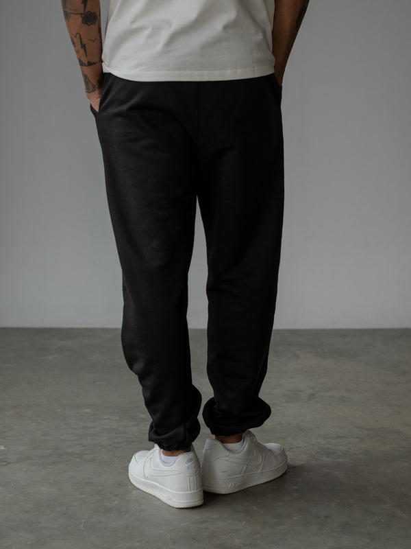 Vista posterior de modelo de pie, usando jogger color negro con bota elástica y camiseta oversized crudo.