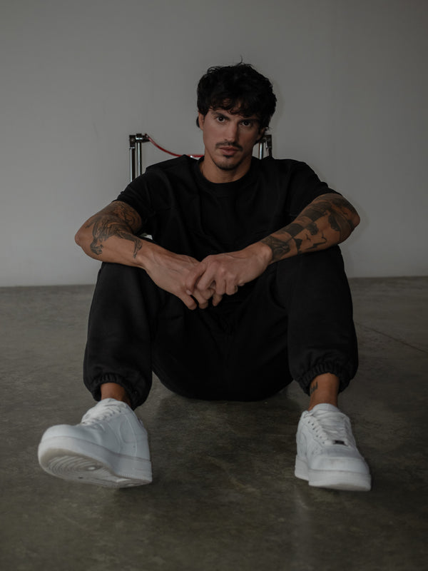 Modelo sentado en el suelo con las manos juntas, usando jogger color negro con bota elástica y camiseta oversized negra.