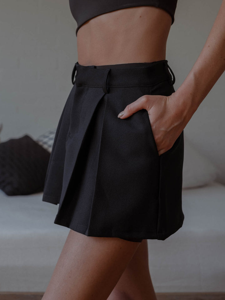 Vista lateral de mini falda color negro con pliegues frontales y bolsillo lateral.