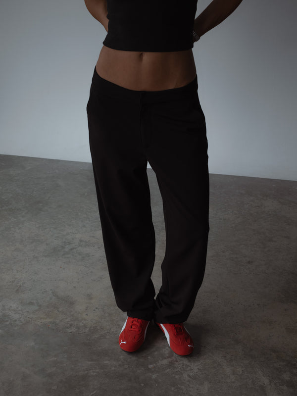 Plano medio de modelo con las manos en las caderas, mostrando un pantalón clásico color negro de tiro medio y bota recta, combinado con un top asimétrico color negro.