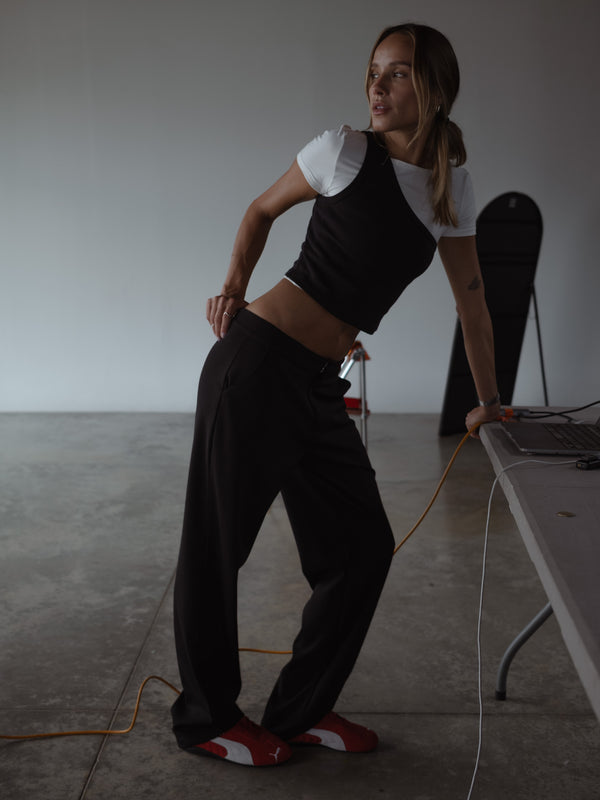 Modelo inclinándose sobre una mesa, usando una camiseta color crudo bajo un top asimétrico color negro y un pantalón color negro con bota recta.