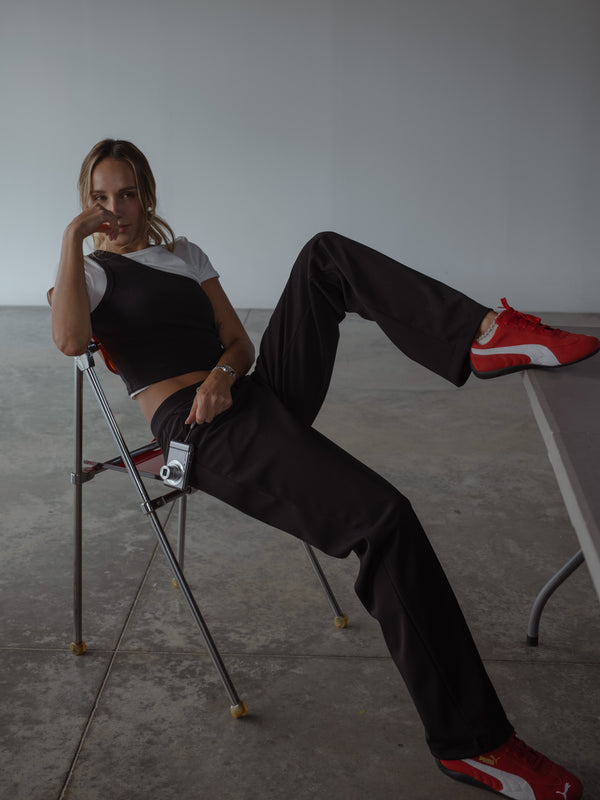 Modelo sentada en una silla, usando un pantalón negro clásico de tiro medio y bota recta, una camiseta manga corta color crudo y un top asimétrico color negro.