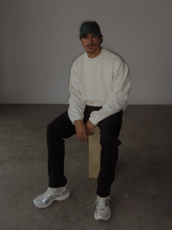 Modelo sentado, usando una sudadera color negro, un buzo oversized color crema y una gorra verde.