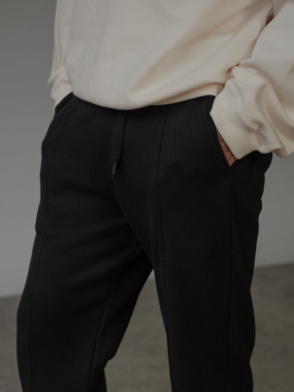 Primer plano de un pantalón color negro y un buzo color crema. El modelo tiene una mano dentro del bolsillo del pantalón.