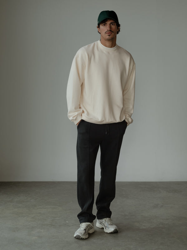 Modelo de pie con las manos en los bolsillos, usando un pantalón color negro, una gorra verde y un buzo color crema.