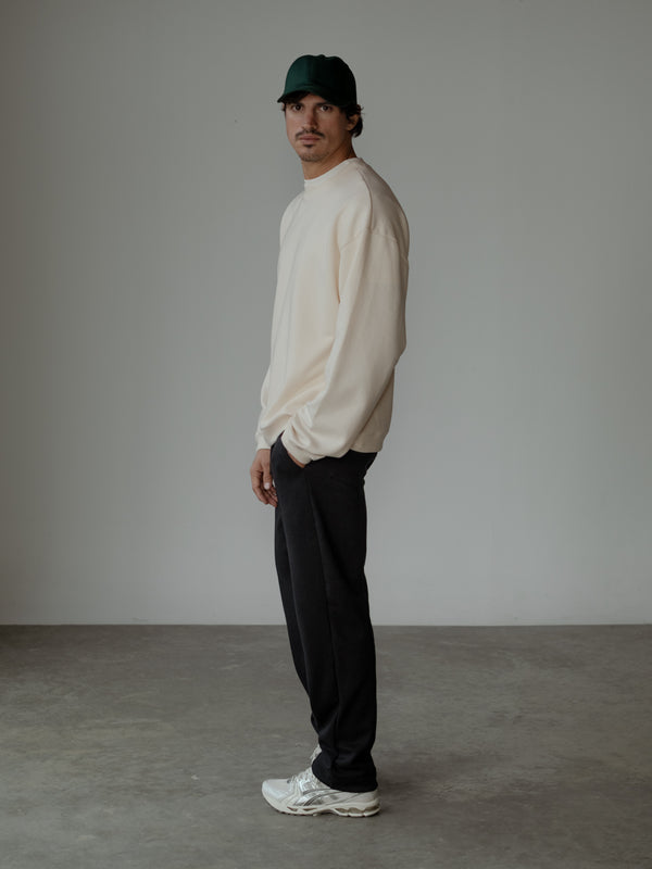 Modelo de perfil izquierdo, usando una gorra verde, un buzo oversized color crema y un pantalón color negro.