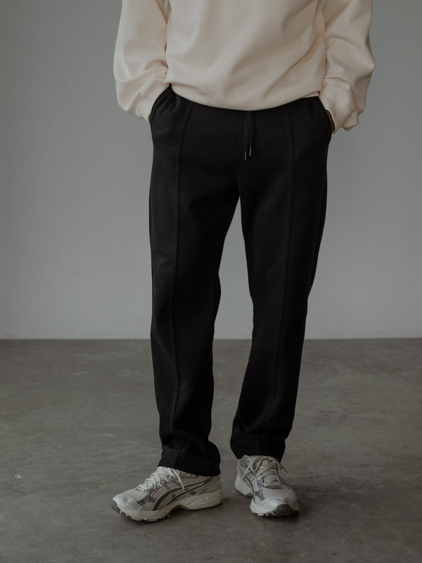 Modelo de pie con las manos en los bolsillos, usando un buzo color crema y un pantalón color negro con cortes frontales.