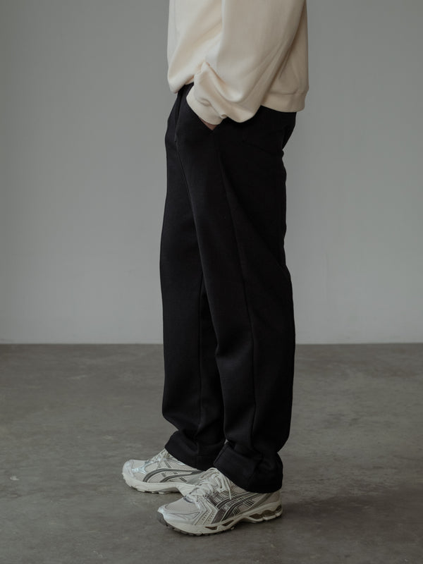 Modelo de perfil izquierdo con las manos en los bolsillos, usando un buzo color crema y un pantalón de sudadera color negro.