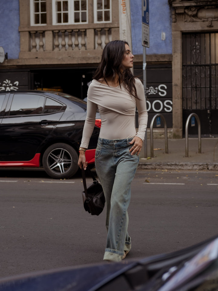 Mujer cruzando la calle mientras sostiene un bolso de mano color cafe,un body color crema y un pantalon de denim.