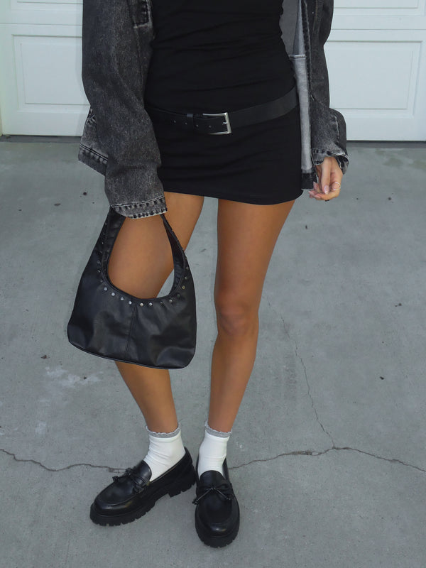 Modelo sosteniendo mini bolso negro usando vestido negro y chaqueta.